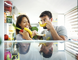 Two happy young adults holding produce in front of a refrigerator.